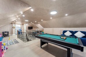 A pool table with a green felt surface, a blue sofa with white cushions, and a ping pong table in a room with a sloped ceiling and recessed lighting.