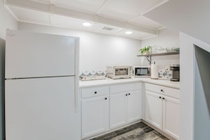 Una cocina con armarios blancos, un refrigerador, un microondas, una cafetera y una repisa con plantas.