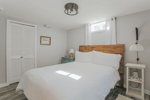 Un dormitorio con cama, mesita de noche, lámpara, ventana con cortinas y puerta con contraventanas.