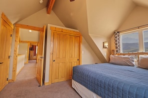 Une chambre avec un plafond en pente, des portes en bois, un lit recouvert d’une courtepointe bleue et une fenêtre donnant sur un beau paysage.