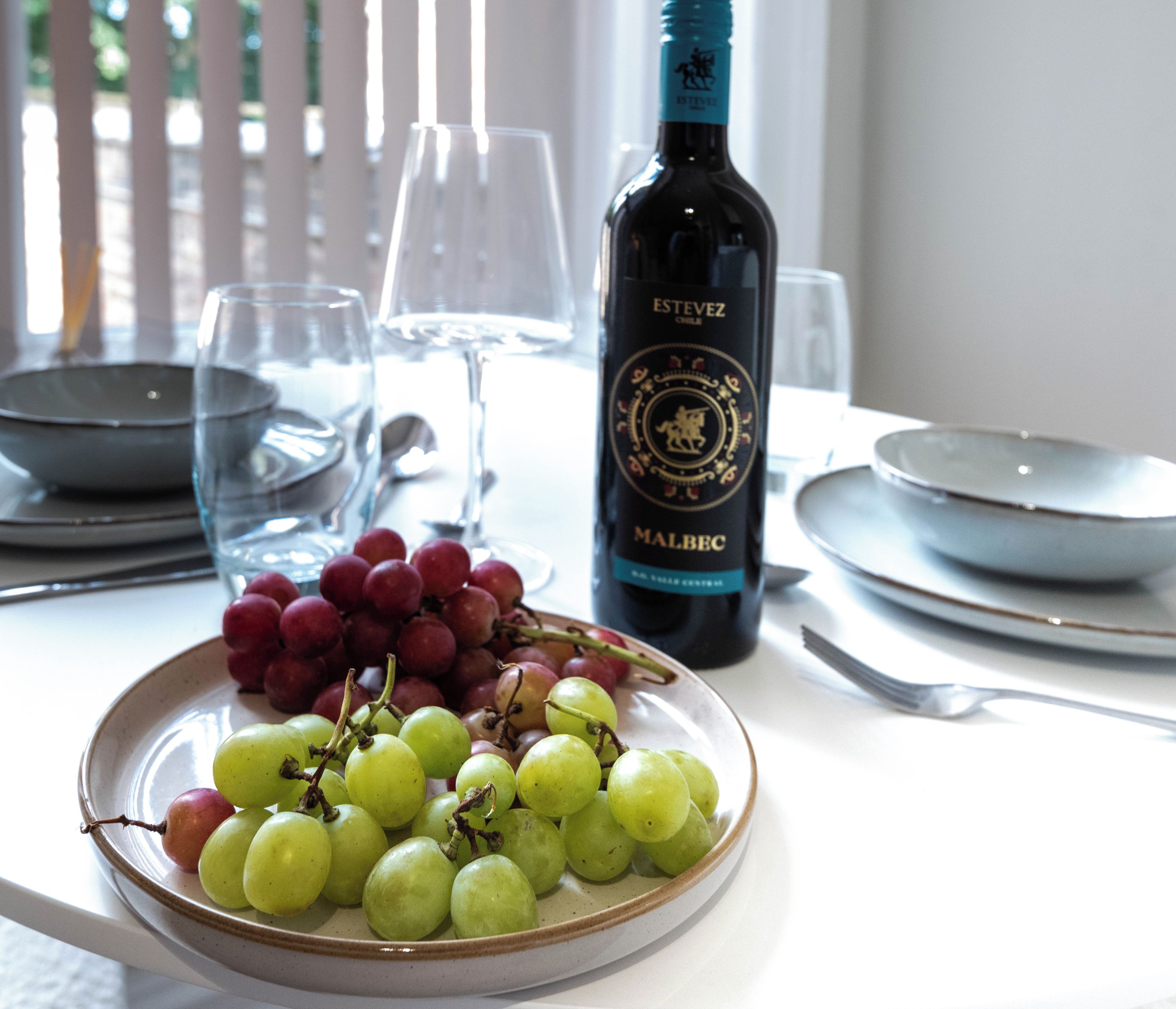 Une table à manger avec une assiette de raisins, une bouteille de vin et des verres vides.