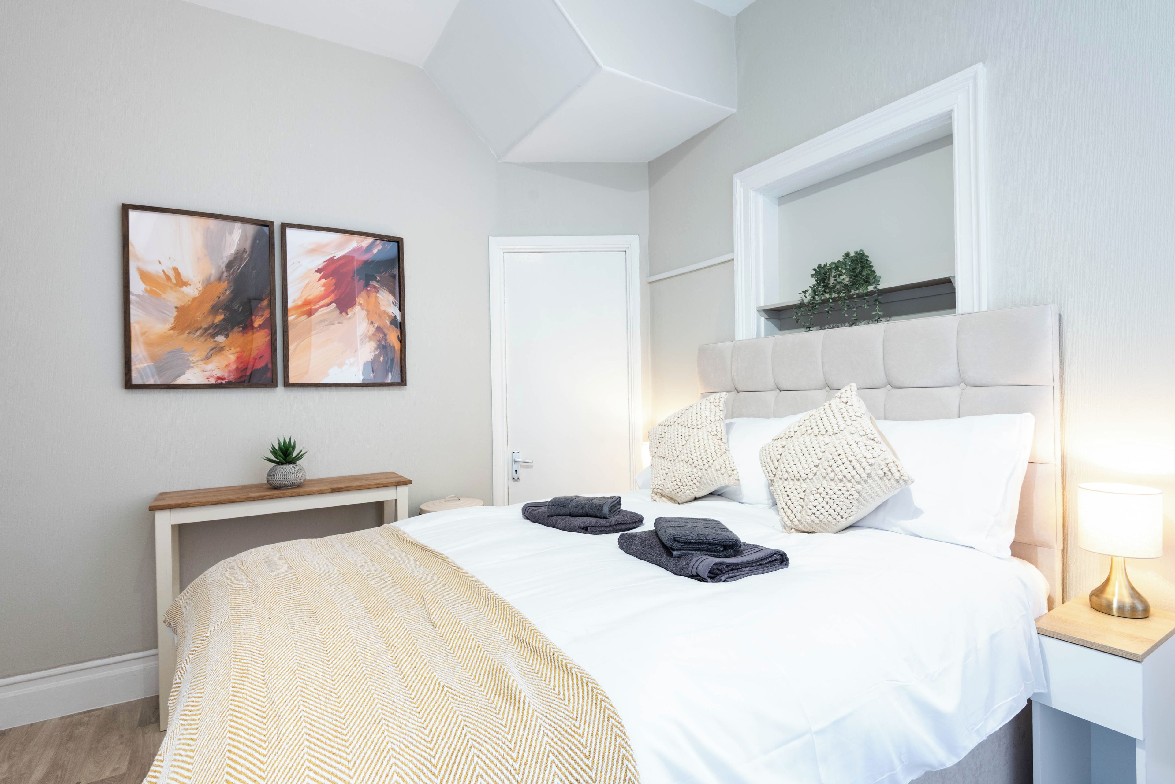 A hotel room with a bed, a bedside table, a mirror, and two abstract paintings on the wall.