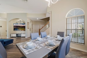 A dining area with a long table set for four, a staircase, and a large window with vertical blinds. - Bright & Spacious 6BR Near Disney w/Pool (Davenport)