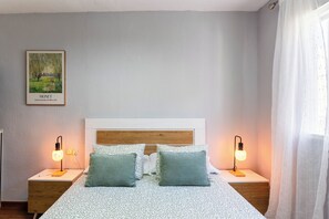 Ein Schlafzimmer mit einem Bett, zwei Nachttischlampen, einem gerahmten Gemälde und einem Fenster mit Vorhängen.
