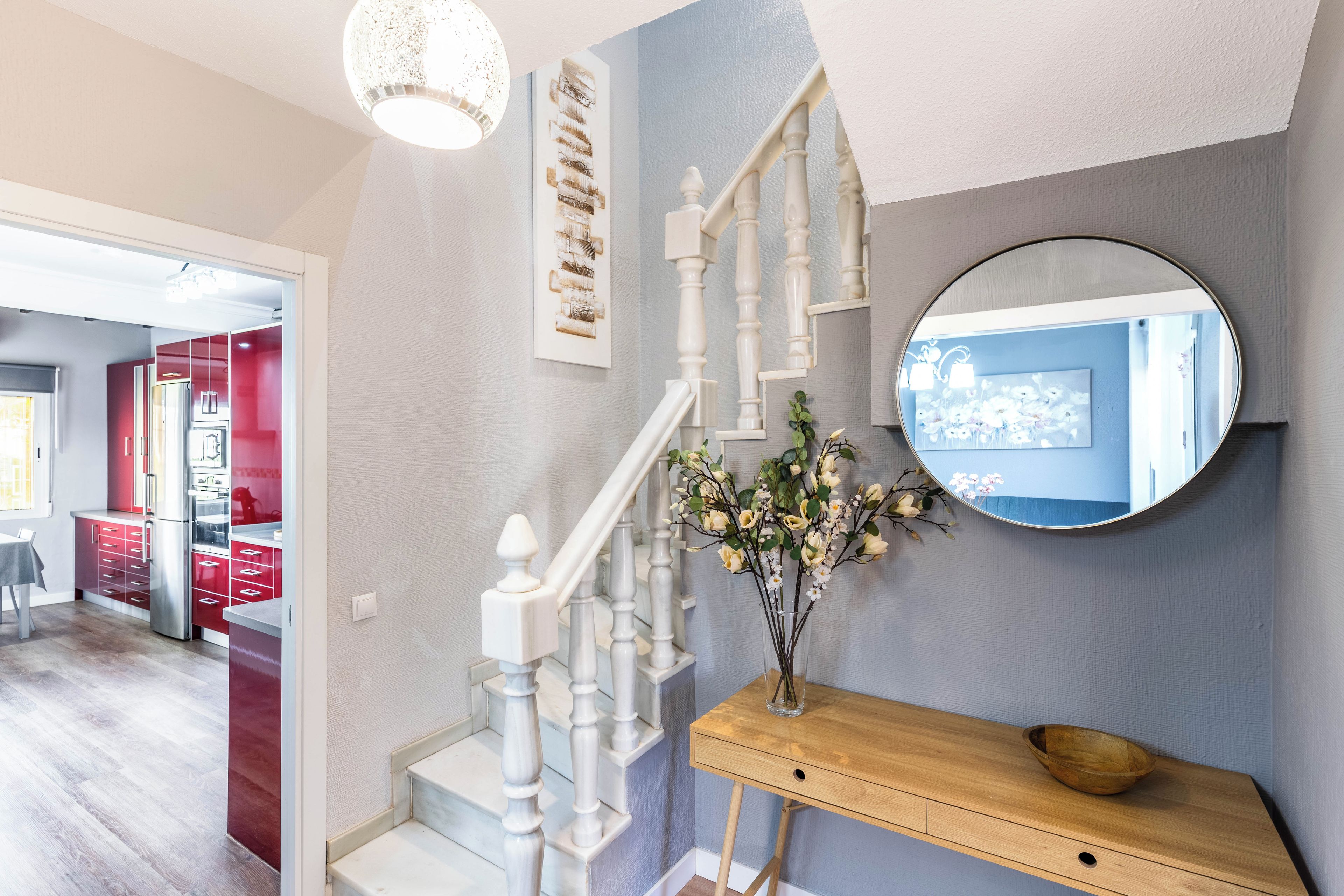 Een trap met witte balustrades, een houten bijzettafeltje met een bloevas, en een ronde spiegel aan de muur.
