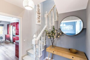 Een trap met witte balustrades, een houten bijzettafeltje met een bloevas, en een ronde spiegel aan de muur.