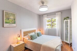 Een slaapkamer met een bed, nachtkastjes, een raam met gordijnen, een plafondventilator en een schilderij in een lijst aan de muur.
