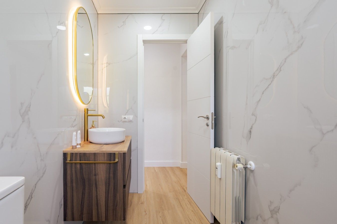Un baño moderno con pared de mármol, tocador de madera, lavamanos blanco y espejo con marco dorado.