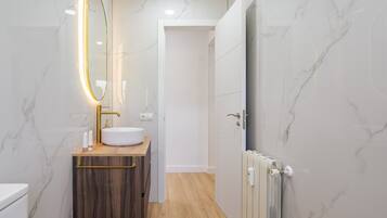 Un baño moderno con pared de mármol, tocador de madera, lavamanos blanco y espejo con marco dorado.