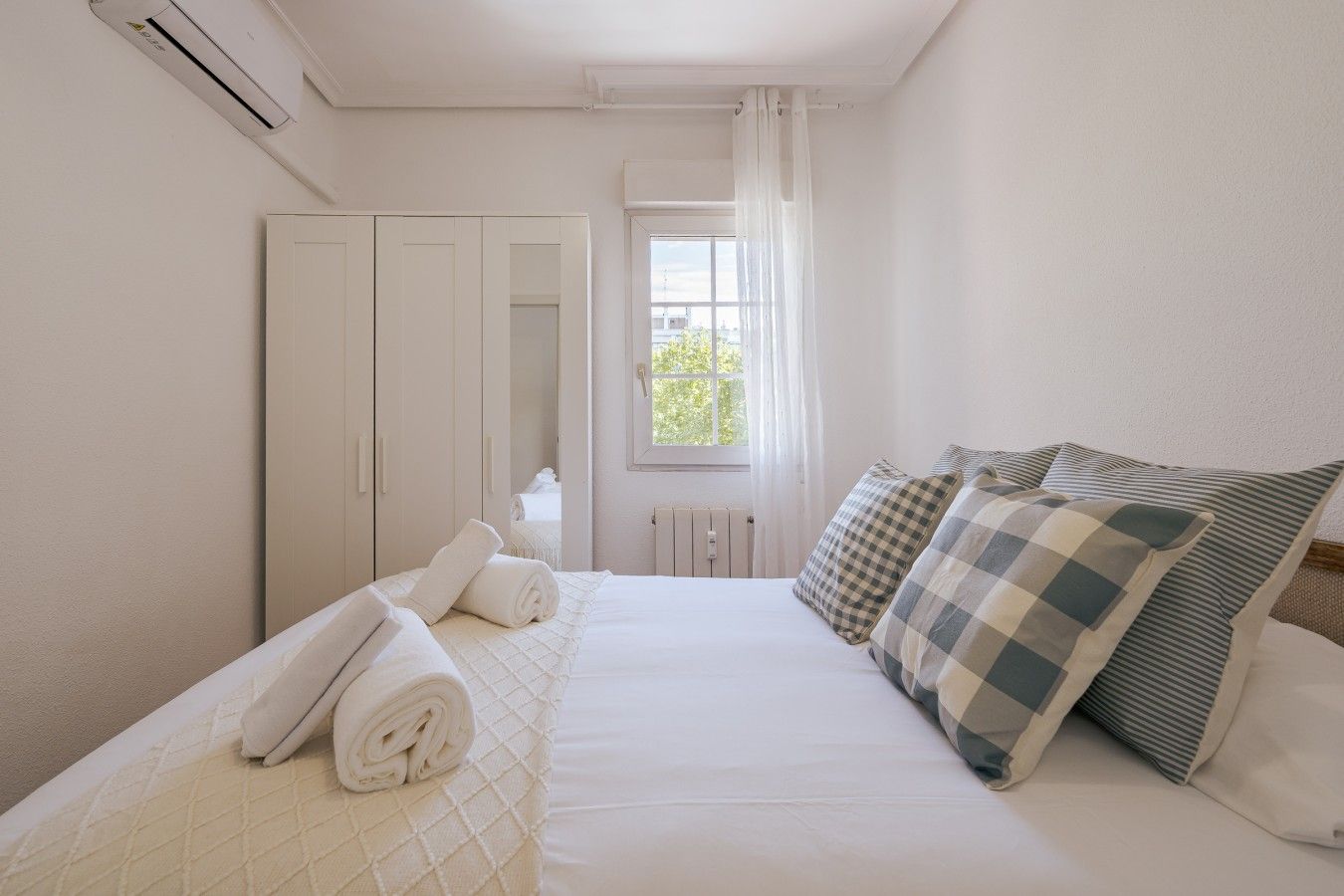 Un dormitorio con cama, almohadas, una toalla enrollada, un armario, una ventana con cortinas y un equipo de aire acondicionado.