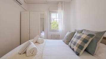 Un dormitorio con cama, almohadas, una toalla enrollada, un armario, una ventana con cortinas y un equipo de aire acondicionado.