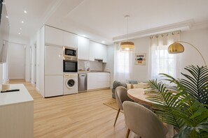 Una cocina moderna con electrodomésticos integrados, un comedor con mesa y sillas redondas, y una sala de estar con sofá y una planta.