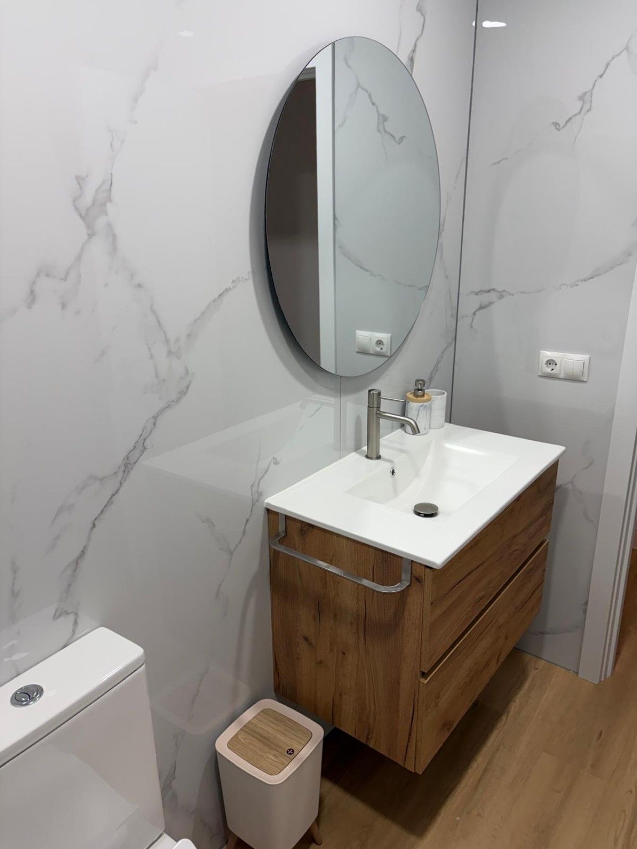 Un baño con pared de mármol blanco, un espejo redondo, un lavabo blanco, una cómoda de madera y un cubo de basura blanco.