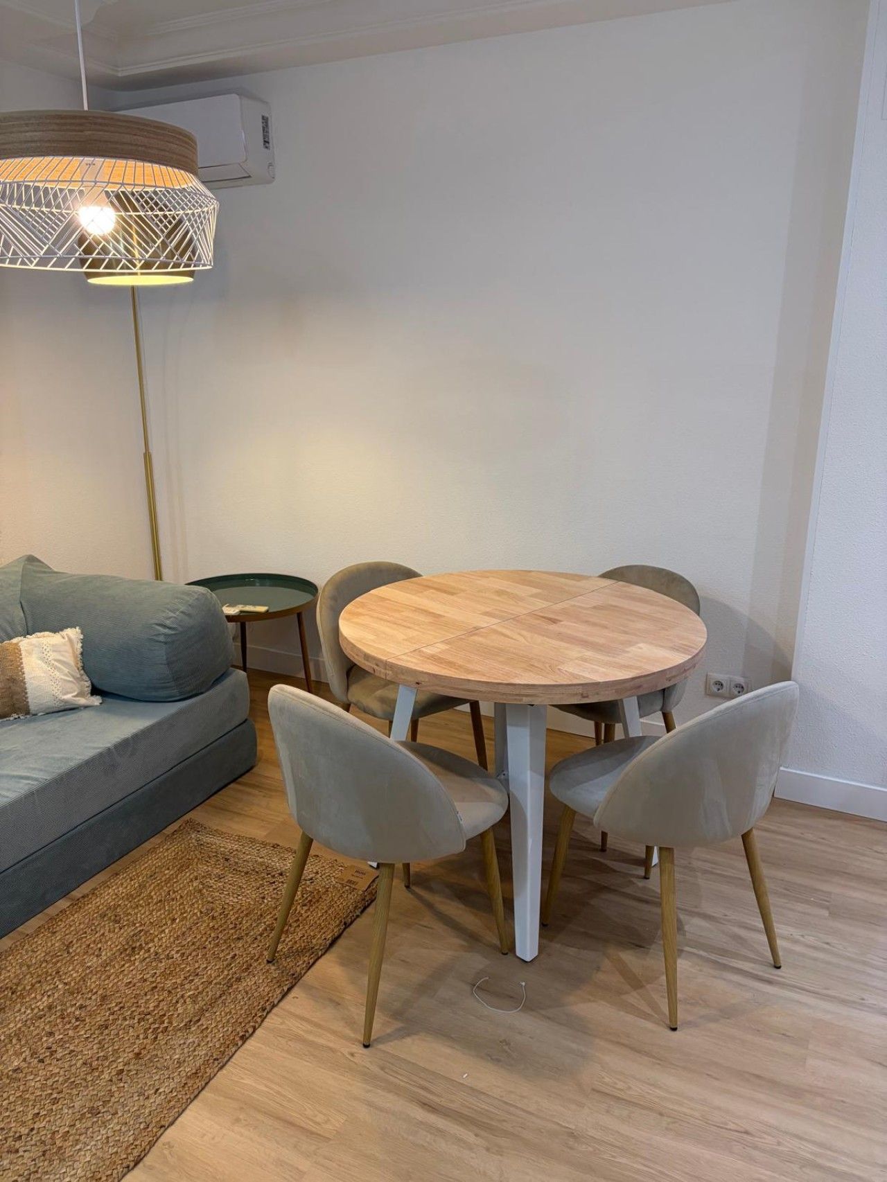 Una sala de estar moderna con una mesa redonda de madera, sillas tapizadas en gris, un sofá gris con cojines, un tapete tejido y una lámpara colgante.