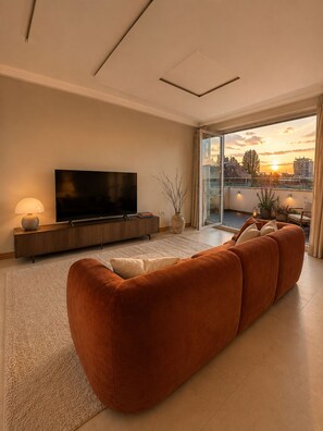 A modern living room with a large flat-screen TV, a sofa, and a view of the sunset through a glass door. - Luxury Marylebone Penthouse (London)