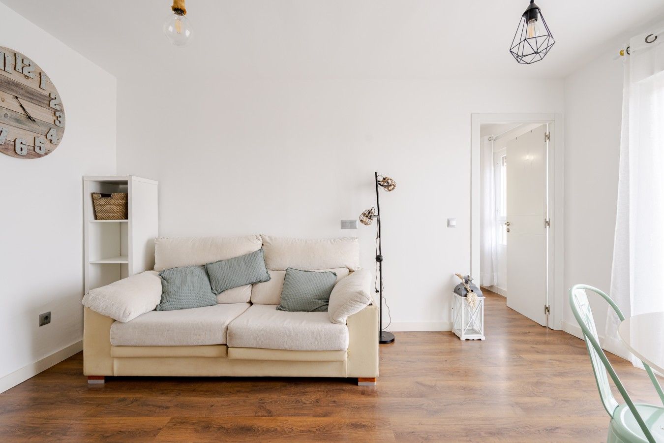 Una sala de estar con un sofá color beige, una estantería blanca, una lámpara de pie y una puerta que da a otra habitación.