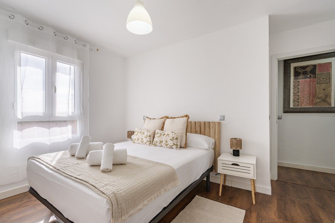 A bedroom with a bed, bedside tables, a nightstand, a lamp, a window with curtains, and a wall with a decorative piece.