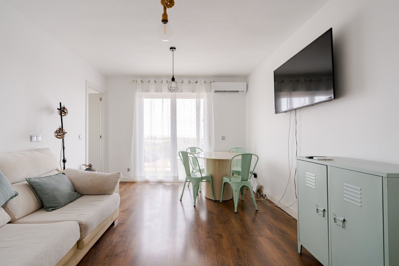Una sala de estar con sofá, una mesa de comedor con sillas, un televisor montado en la pared y un mueble.