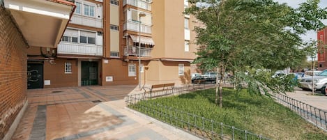 A multi-story residential building with a paved courtyard, a small tree, and a metal fence.