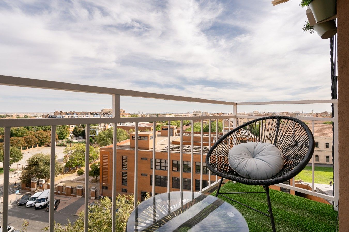 Un balcón con una silla de mimbre negro y una mesa de vidrio, con vista a un paisaje urbano que incluye edificios y un área verde.