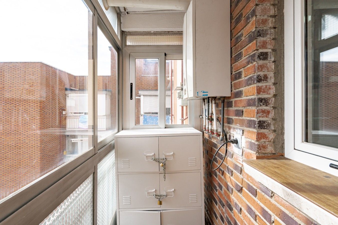 A narrow corridor with a brick wall, a window, and a white cabinet with a lock.