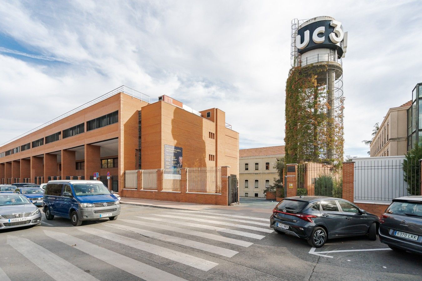 Un estacionamiento con varios coches, un edificio con un letrero que dice 'JC3' y una estructura alta cubierta de hiedra.