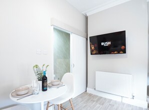 Un coin repas moderne avec une table ronde, des chaises blanches, un saladier, des verres à vin et une bouteille. Un téléviseur à écran plat est fixé au mur.