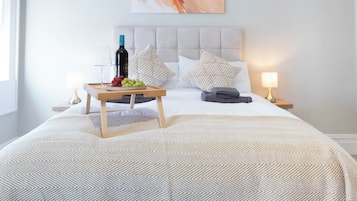 Un lit bien fait, recouvert d’une couvre-lit à motifs, une table de chevet avec une bouteille et des raisins, et deux lampes sur des tables de nuit.