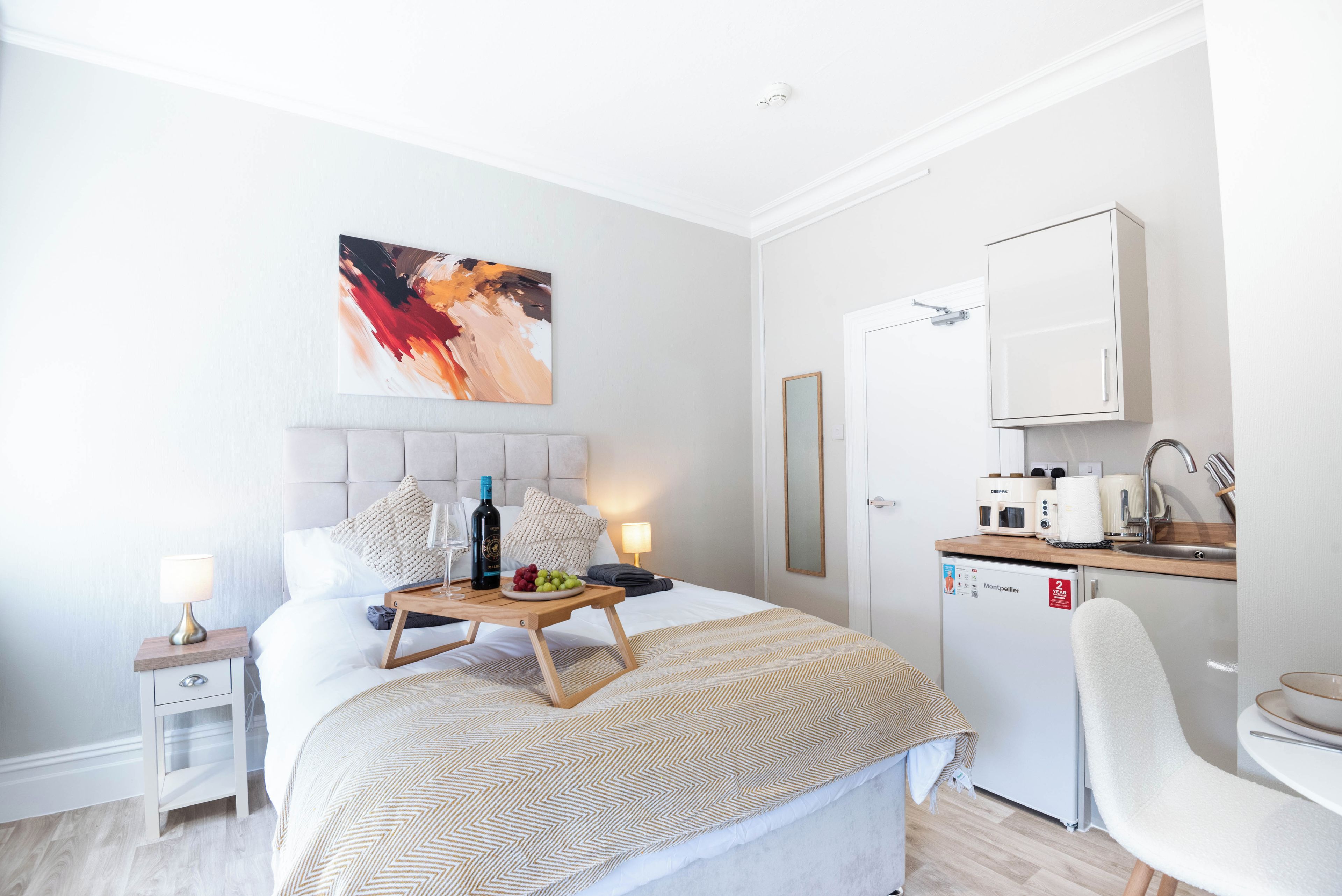 A hotel room with a bed, bedside table, lamp, artwork, and a kitchenette with a refrigerator, microwave, and coffee maker.