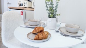 Ein runder Esstisch mit einem weißen Stuhl, zwei Schüsseln, zwei Weingläsern, einer Topfpflanze und zwei Croissants auf einem Teller.