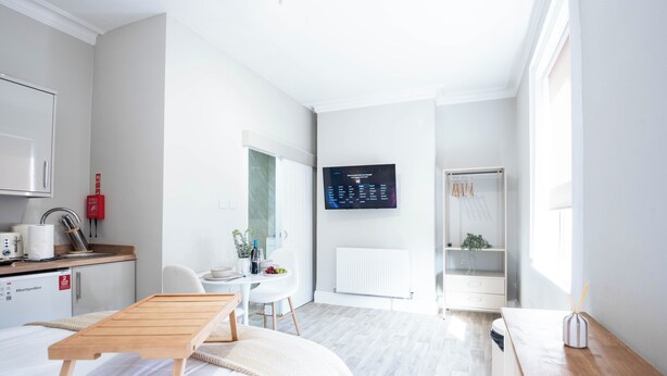 Ein modernes Hotelzimmer mit Bett, Schreibtisch, einer kleinen Küchenzeile und einem an der Wand montierten Fernseher.