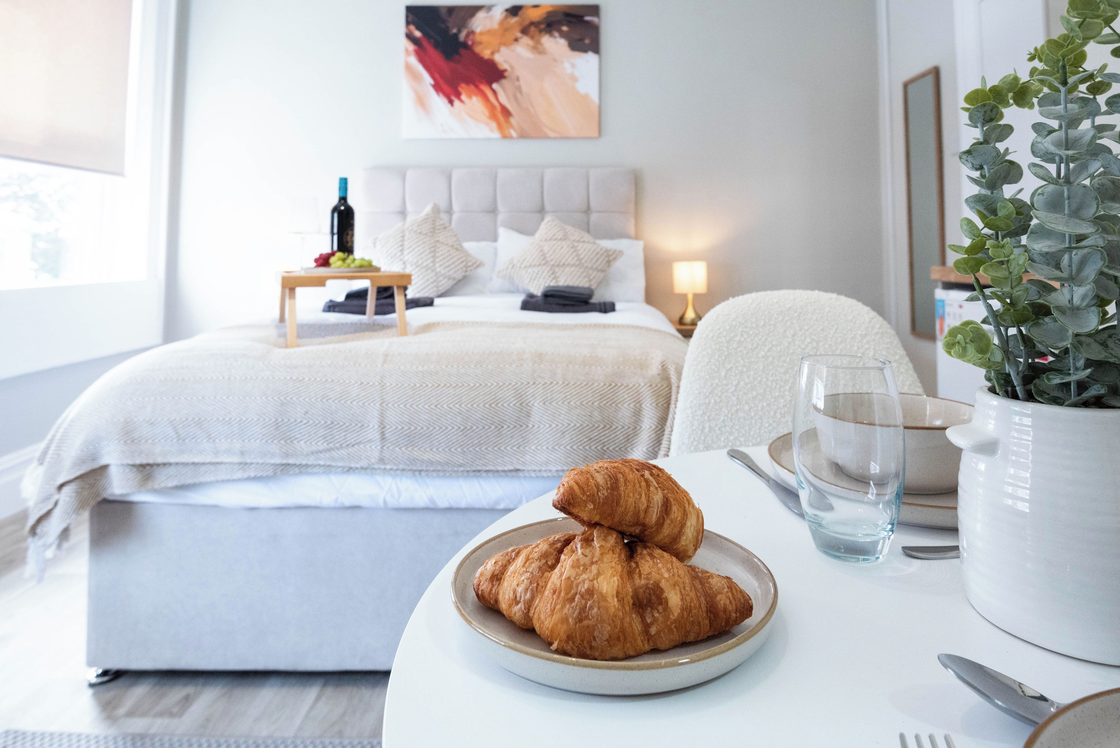 A neatly arranged hotel room with a bed, bedside table with a bottle, a plate with croissants, a glass of water, and a potted plant.