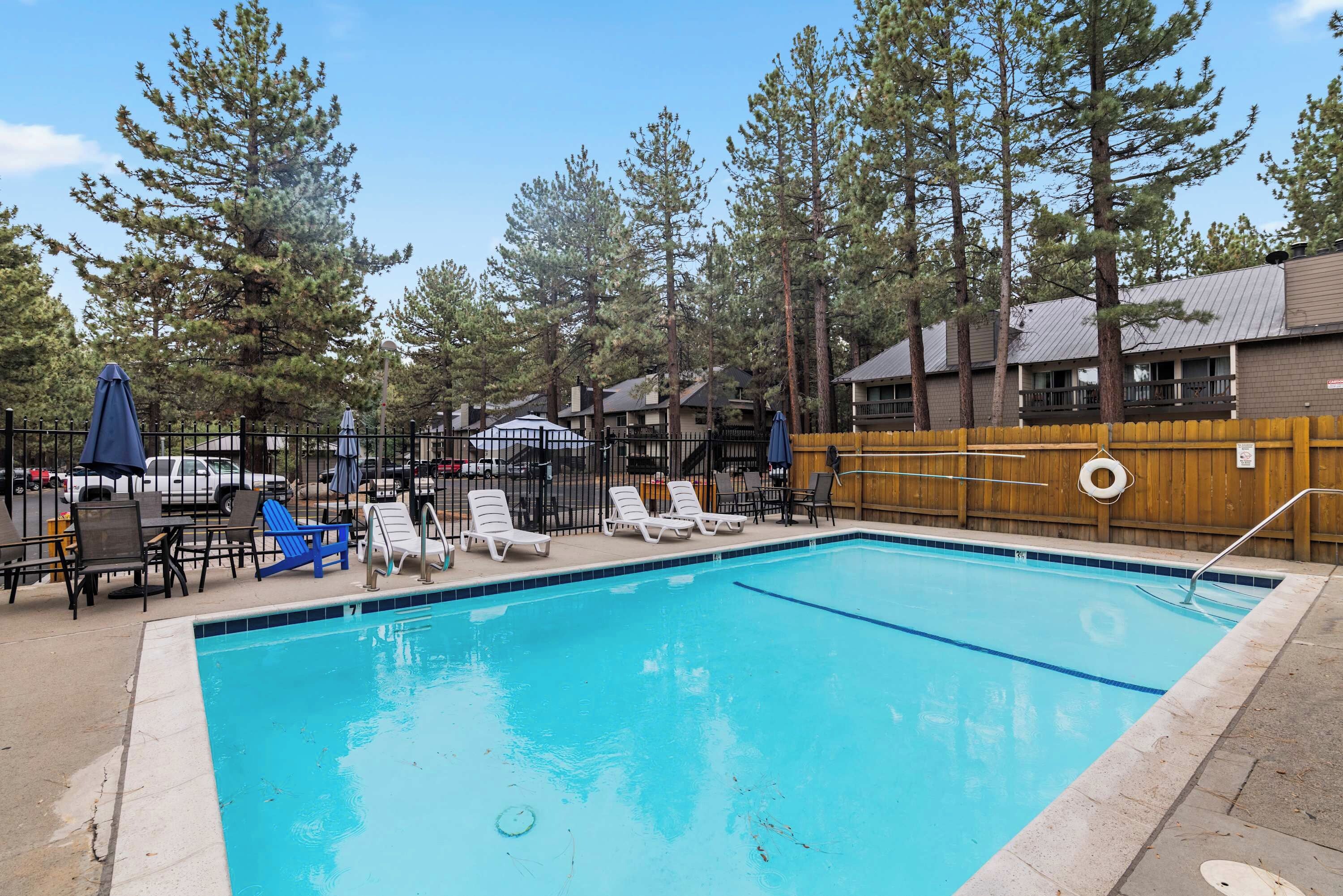 A rectangular swimming pool with clear blue water, surrounded by a wooden fence, and equipped with lounge chairs and umbrellas.