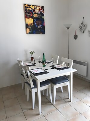 Un coin repas avec une table blanche, quatre chaises, une plante en pot, des verres à vin et une bouteille.