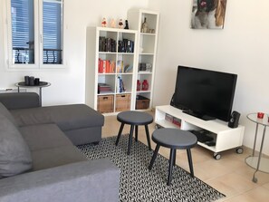 A modern living room with a grey sectional sofa, a flat-screen TV on a white stand, a small round table, and a bookshelf filled with books and decorative items. - Holiday Home 'Le Rêve D'enfant' with Private Pool, WiFi, and Air Conditioning (Narbonne)