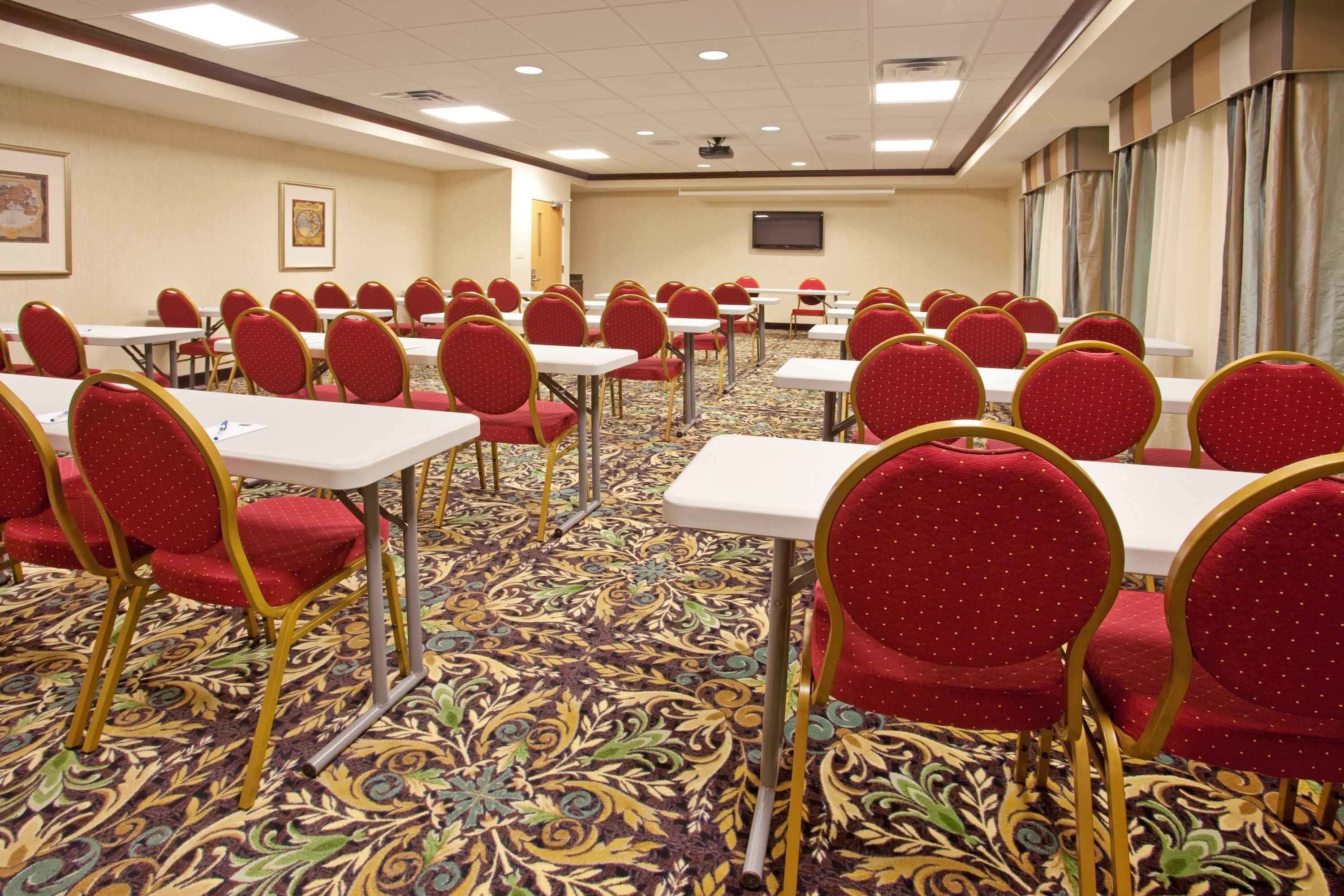 A conference room with rows of tables and chairs, carpeted floor, and a projector screen on the wall.