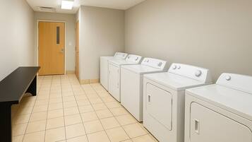 A laundry room with four white washing machines, a bench, and a door at the end of the hallway.