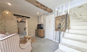 Une pièce avec un mur en pierre, un escalier en bois, un téléviseur posé sur un meuble et un canapé à rayures.