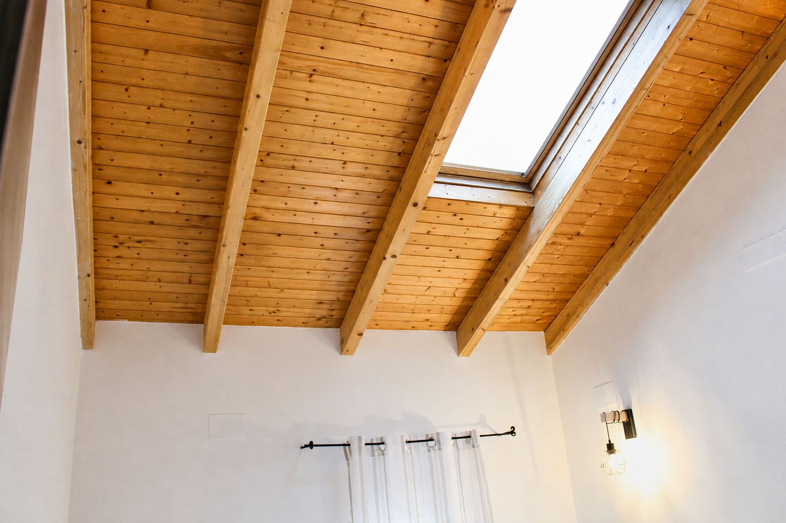 Habitació amb sostre de fusta inclinat, un fanal i una barra de cortina blanca, juntament amb una làmpada fixada a la paret.