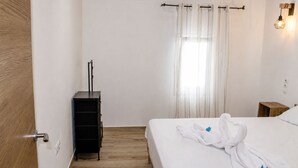 Quarto com piso de madeira, cama com roupa de cama branca, cômoda preta, janela com cortinas leves e luminária fixada na parede.