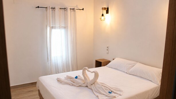 Quarto de hotel com cama, mesa de cabeceira, luminária na parede e cortinas translúcidas.