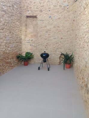 Un patio de paredes de piedra con una parrilla portátil, dos plantas en macetas y luces colgantes.