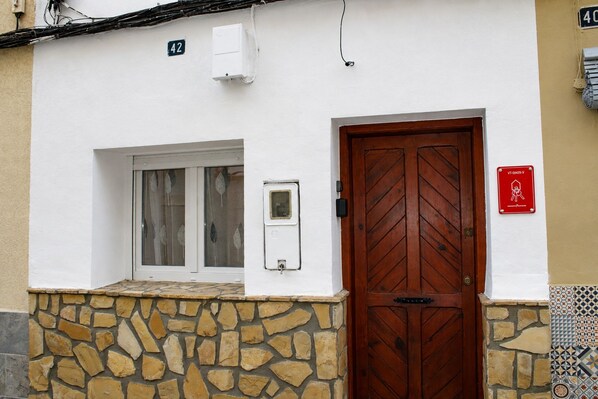 A building with a stone base, a wooden door, a window with curtains, and a number sign above the door. - Casa El Calvario, where you can take a break, relax, and enjoy time with family and friends. (Bolbaite)