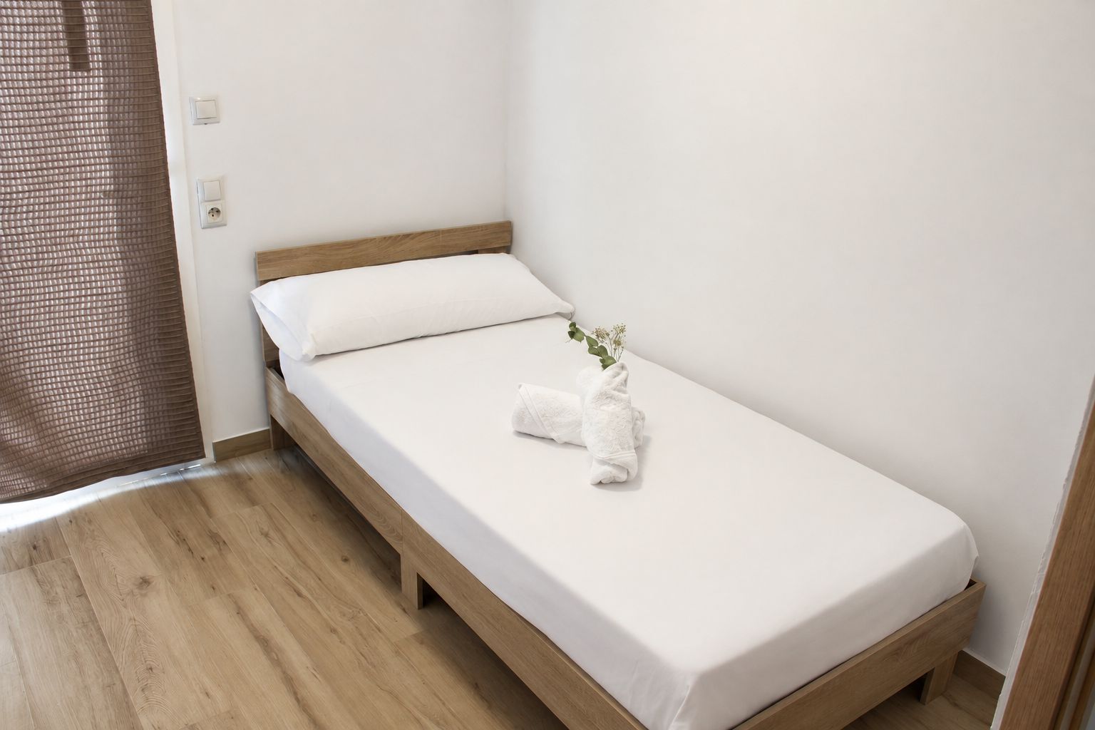 A single bed with a wooden frame and white bedding, a small plant on the bed, and a wooden floor.