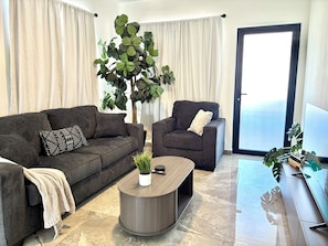 En moderne stue med en grå sofa, en matchende lænestol, et sofabord og en stor plante.