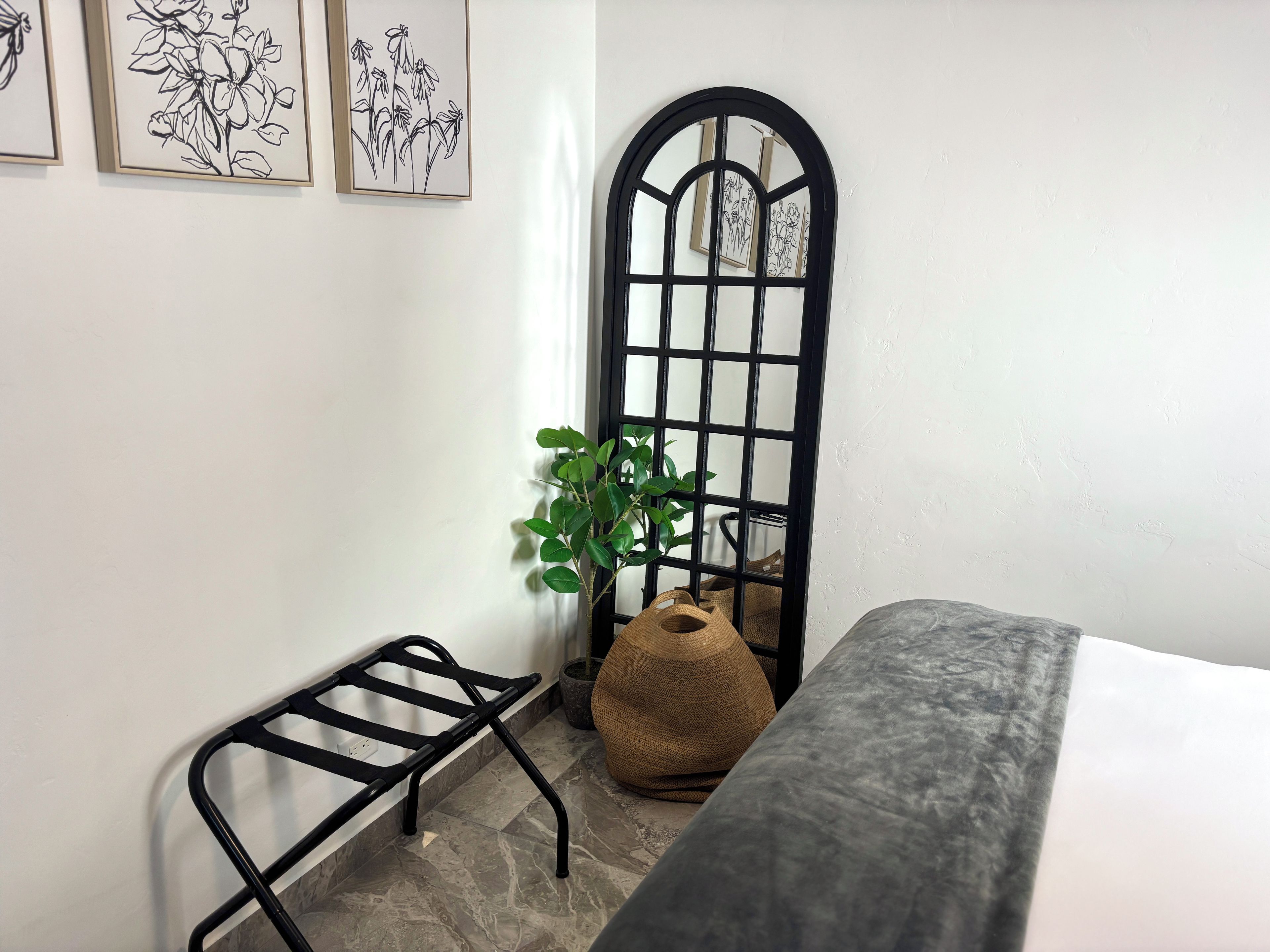 A corner room with a bed, a black metal shelf, a potted plant, and framed artwork on the wall.