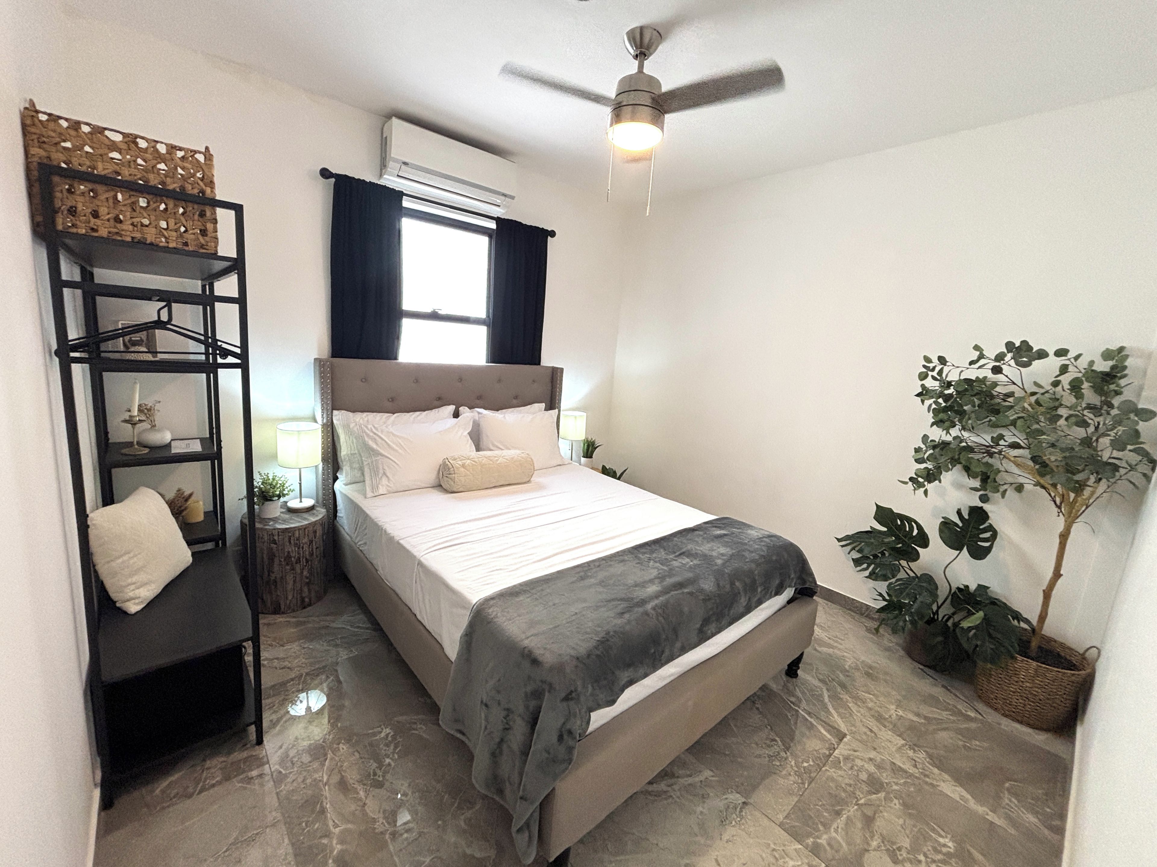 A bedroom with a bed, a ceiling fan, a window with curtains, a wall-mounted air conditioner, a nightstand with a lamp, and a potted plant.