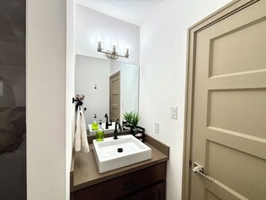 Un baño con lavamanos blanco, un espejo, toallero, una planta y una puerta.