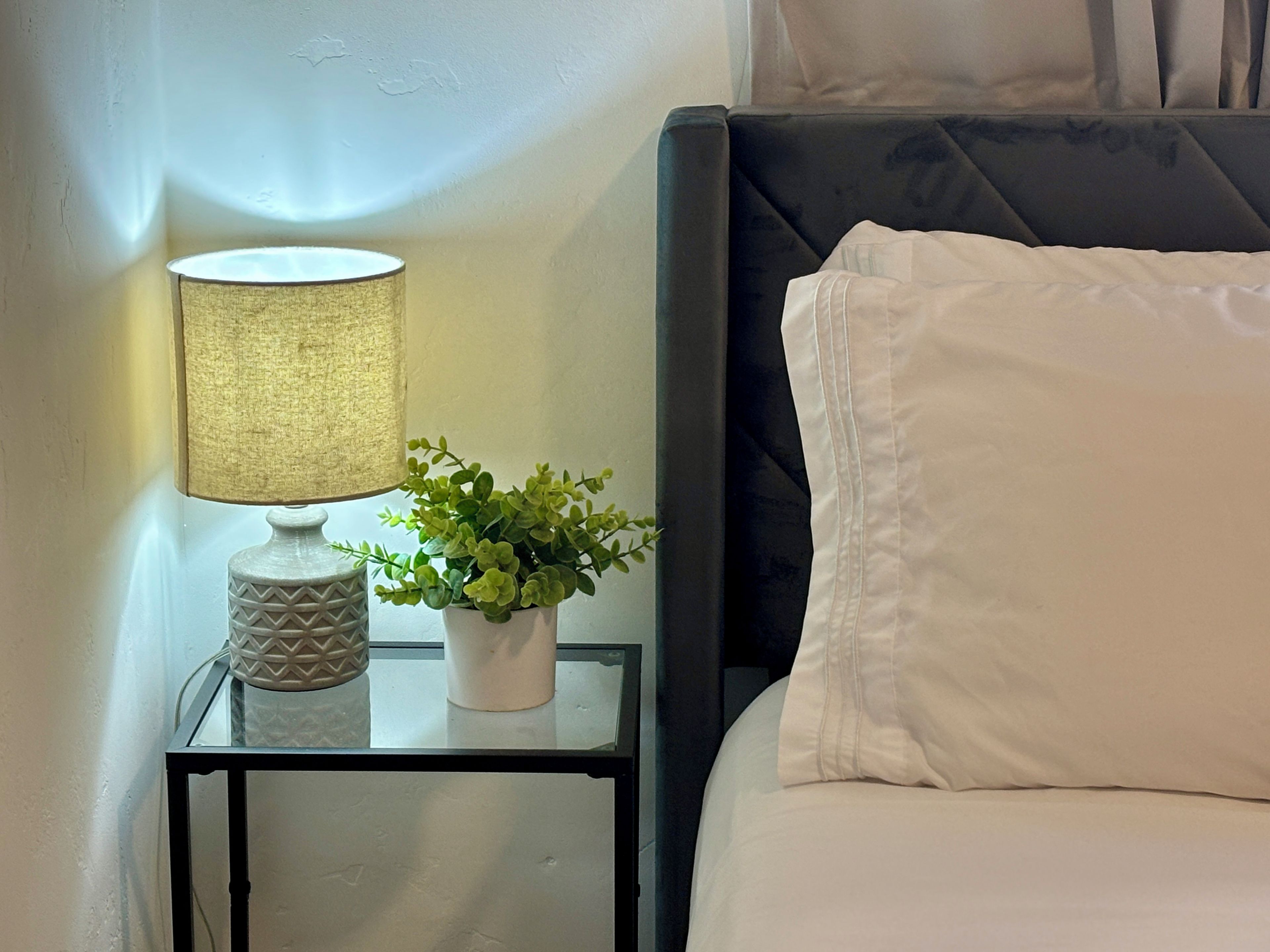 Een net opgemaakt bed met witte beddengoed, een bedlamp met een getextureerde lampenkap, en een kleine potplant op een glazen tafeltje naast het bed.