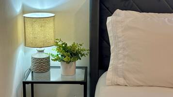 Een net opgemaakt bed met witte beddengoed, een bedlamp met een getextureerde lampenkap, en een kleine potplant op een glazen tafeltje naast het bed.